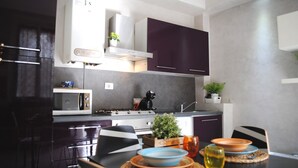 Een moderne keuken met donkere kastjes, een magnetron, een fornuis en een spoelbak. Er staat een eettafel met een plant, een kom en een glas.
