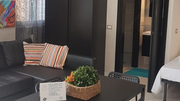 Een moderne woonkamer met een zwarte bank, gestreepte kussens, een kleine tafel met een plant, en een bordje waarop desinfectie- en hygiënematen worden aangegeven.