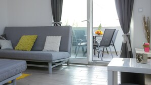 Un saló modern amb un sofà gris, una taula petita i una porta corredissa de vidre que dona a una zona exterior amb una taula i cadires.