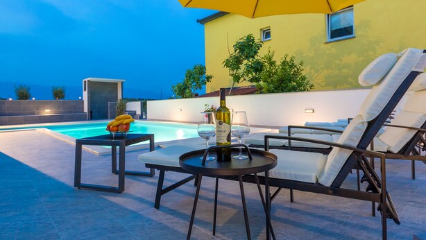 Zona de piscina amb gandules, una taula amb ampolla i vasos, i un parasol groc.