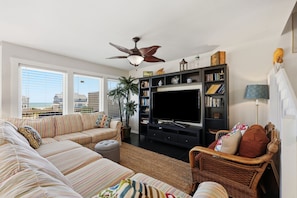 A living room with a sofa, TV, bookshelf, and a ceiling fan. - Ocean View Second-Row Duplex | Spacious 5BR | Beach Access Across Street (Emerald Isle)