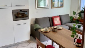 Une cuisine moderne avec des armoires blanches et un four encastré. On y trouve une table à manger avec un chemin de table rouge et blanc, une cafetière et une théière. Un canapé gris avec un coussin rouge est placé contre le mur, et un tableau encadré est accroché au-dessus.