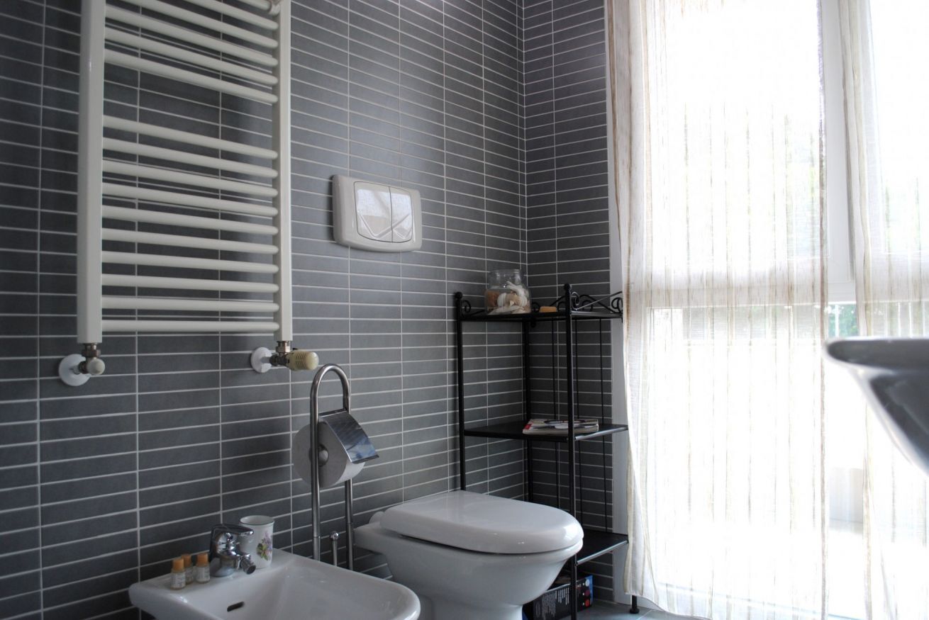 A bathroom with a toilet, sink, and towel rack. There is a window with sheer curtains and a shelf with toiletries.