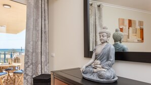 Une statue représentant une figure assise, posée sur une commode, un miroir reflétant une statue de Bouddha et une vue sur la mer par une fenêtre.