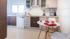 Une cuisine moderne équipée d’un lave-vaisselle, d’une machine à café et d’un mixeur, le tout posé sur le plan de travail. On y trouve également une table à manger avec deux chaises et un set de table rouge.