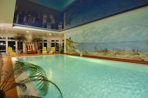 An indoor swimming pool with a mural of a beach scene, lounge chairs, and potted plants.