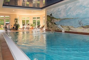 An indoor swimming pool with clear water, surrounded by lounge chairs and potted plants, with a mural of a coastal landscape on the wall. - Aparthotel Am See (Plau am See)