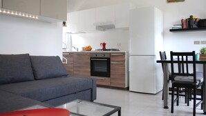 Une cuisine moderne avec des armoires blanches, un four encastré et un réfrigérateur blanc. On y trouve une table à manger avec des chaises et un petit îlot de cuisine sur lequel sont pos és une bouilloire rouge et des oranges.