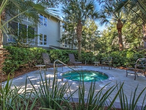 A pool area with lounge chairs and a hot tub surrounded by greenery and a building in the background.