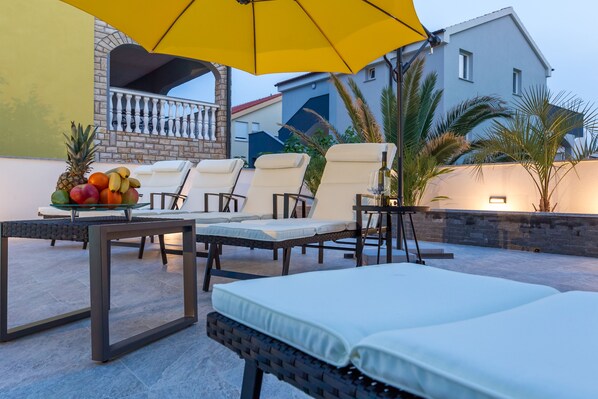Une terrasse avec des chaises longues, une table garnie de fruits et un parasol jaune.