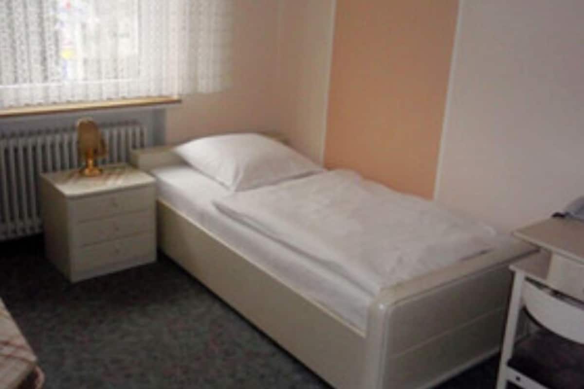 Una habitación pequeña con una cama individual, cabecera de madera, mesita de noche con lámpara y ventana con cortinas traslúcidas.