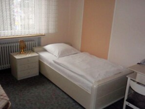 Una habitación pequeña con una cama individual, cabecera de madera, mesita de noche con lámpara y ventana con cortinas traslúcidas.