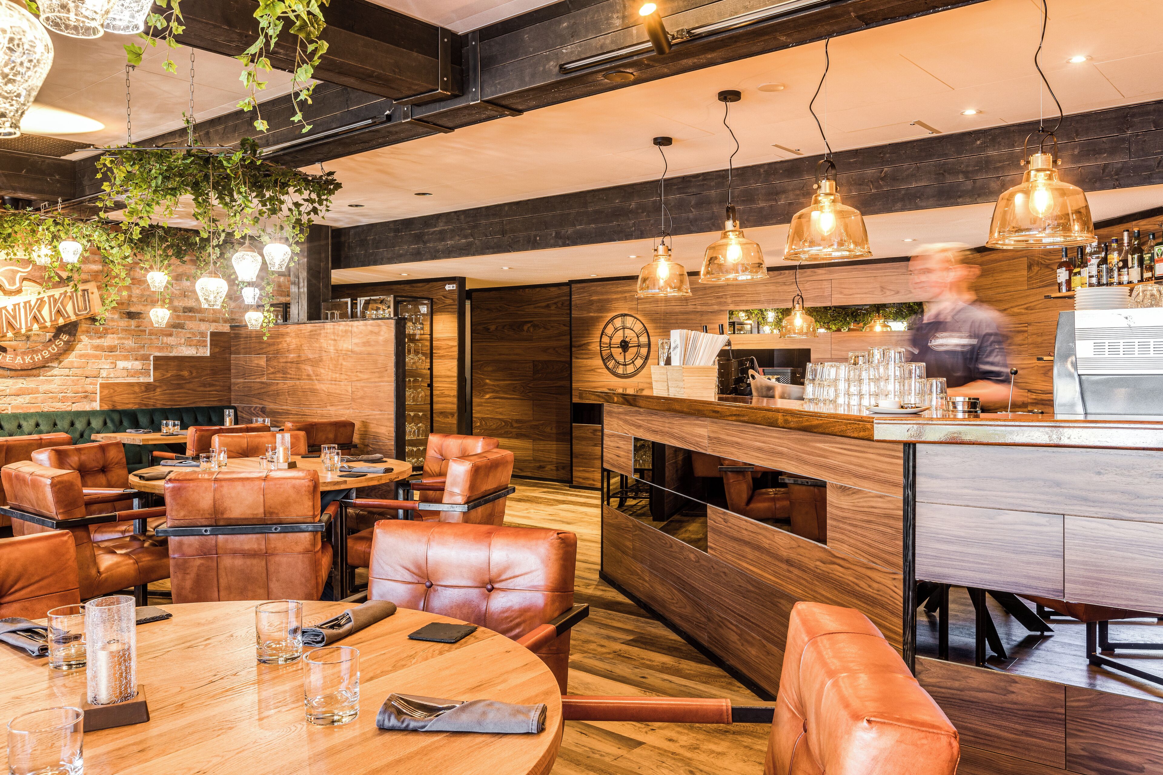 A restaurant with wooden tables and leather chairs, a bar area, and hanging pendant lights.