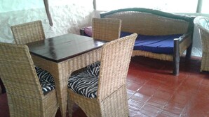 Habitación con una mesa de madera y cuatro sillas de mimbre con cojines estampados de cebra. Se ve una cama con colchón azul y cabecera trenzada.