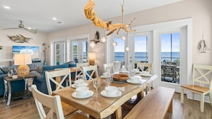 Un coin repas avec une table en bois dressée pour quatre personnes, des chaises blanches et une vue sur la mer grâce à de grandes fenêtres.