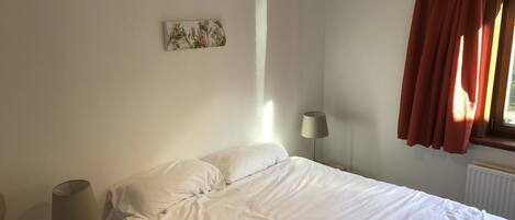 Ein Schlafzimmer mit einem Bett, einer Nachttischlampe, einer Hängelampe, einem Fenster mit Vorhängen und einem Wandbild.