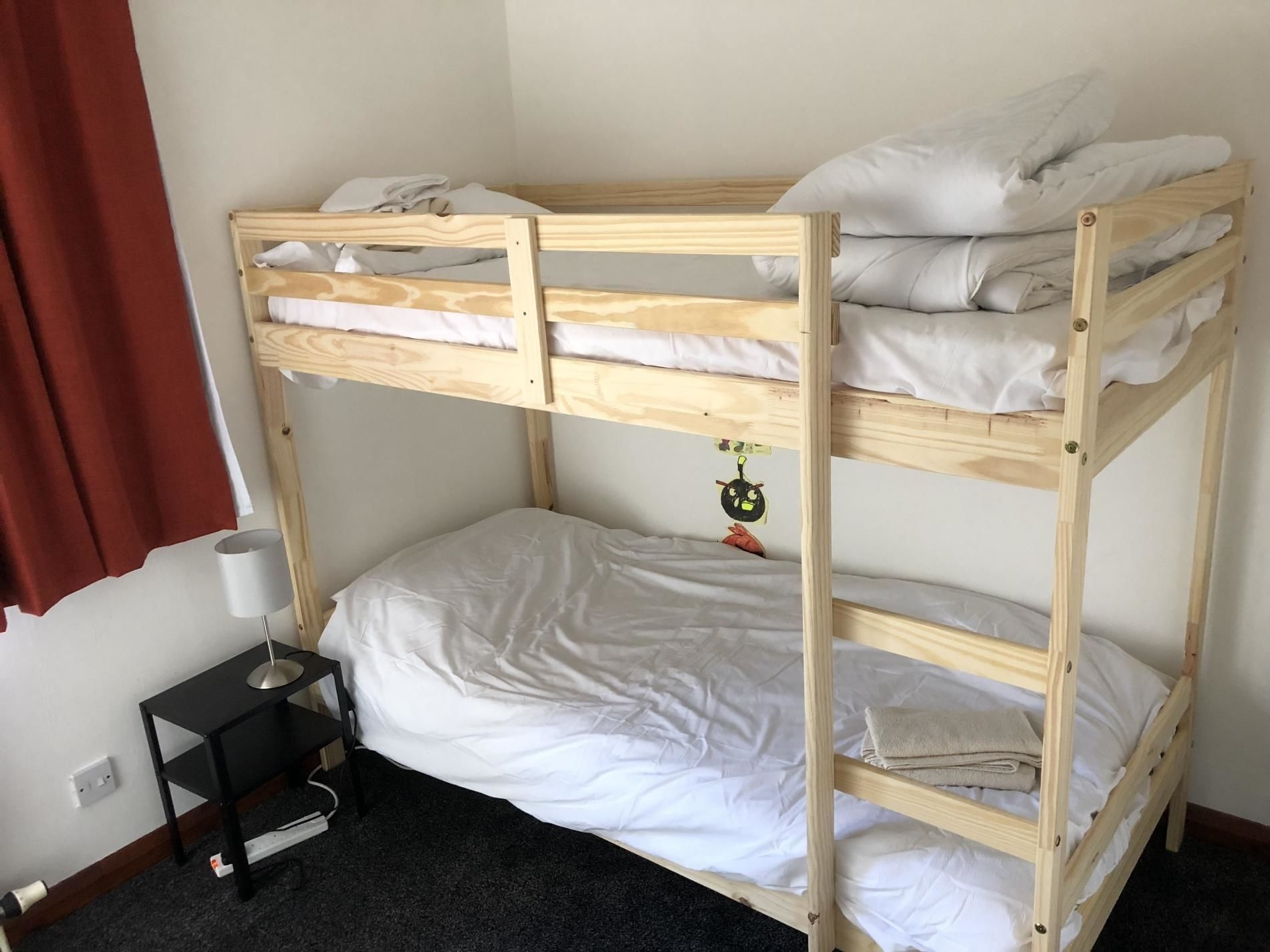 A bunk bed with white bedding, a small bedside table with a lamp, and a red curtain.