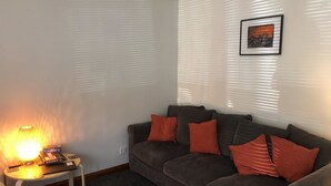 Ett vardagsrum med en grå soffa, röda kuddar, ett litet bord med en lampa och en inramad tavla på väggen.