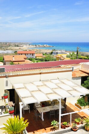 Terraza en la azotea con mesa y sillas, rodeada de plantas en macetas, con vistas a un paisaje costero que incluye edificios y el mar.