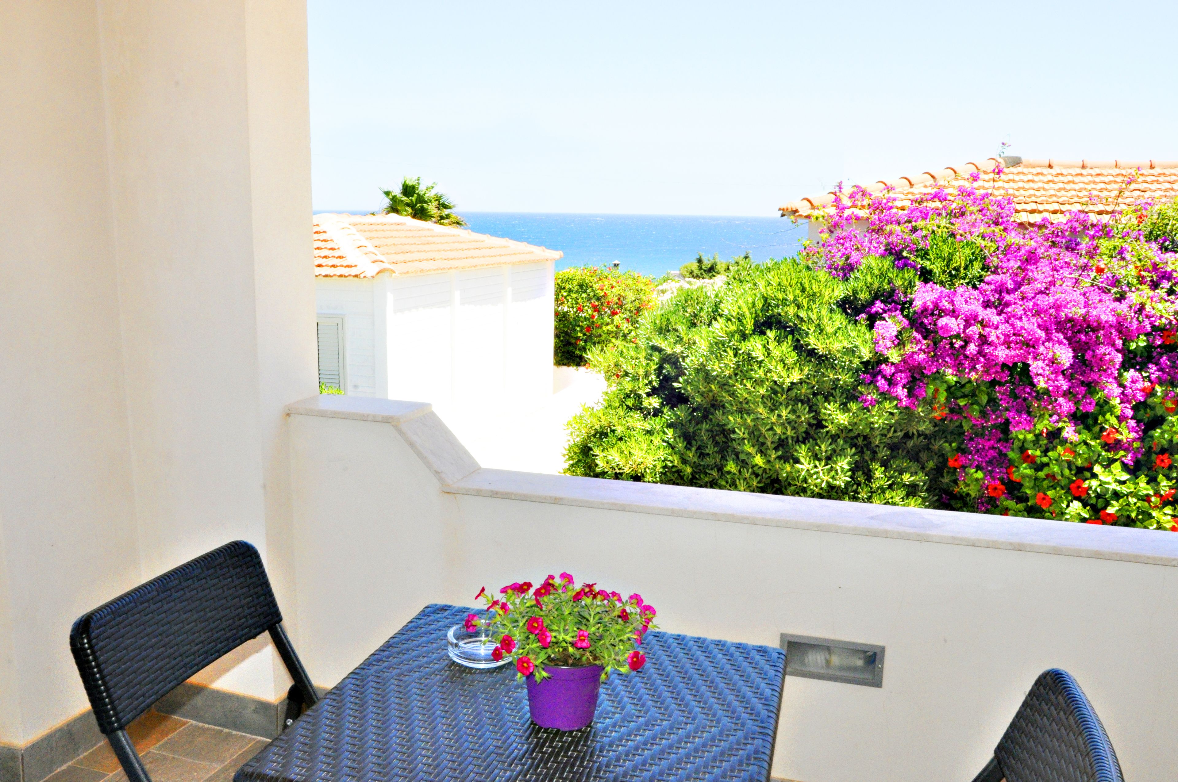 A balcony with a table and chairs, a potted plant, and a view of the sea and buildings.