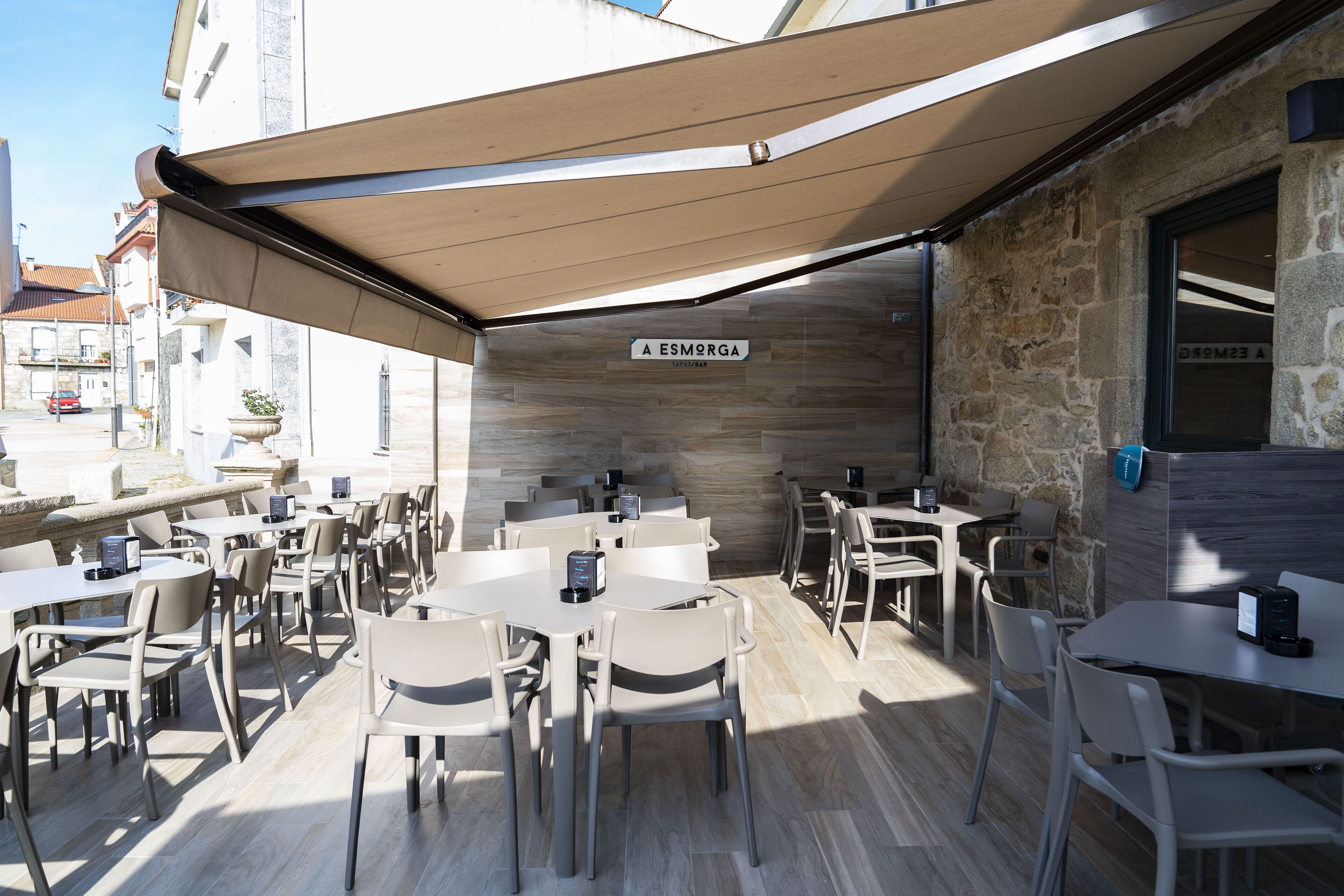 An outdoor cafe area with multiple tables and chairs, a stone wall, and a sign reading "A ESMORGA".