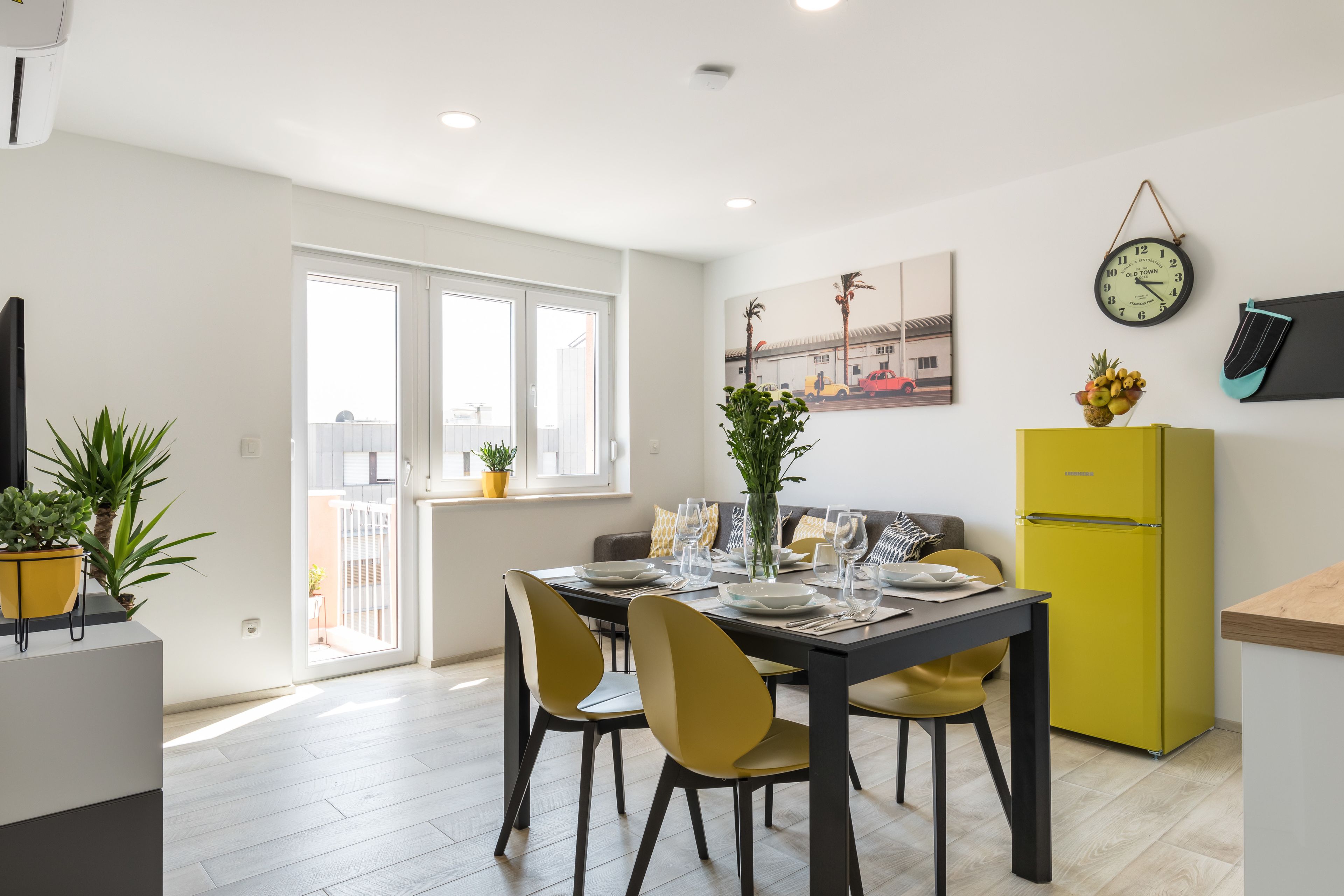 A modern kitchen with a dining area, featuring a yellow refrigerator, a dining table set for four, and a large window providing natural light.