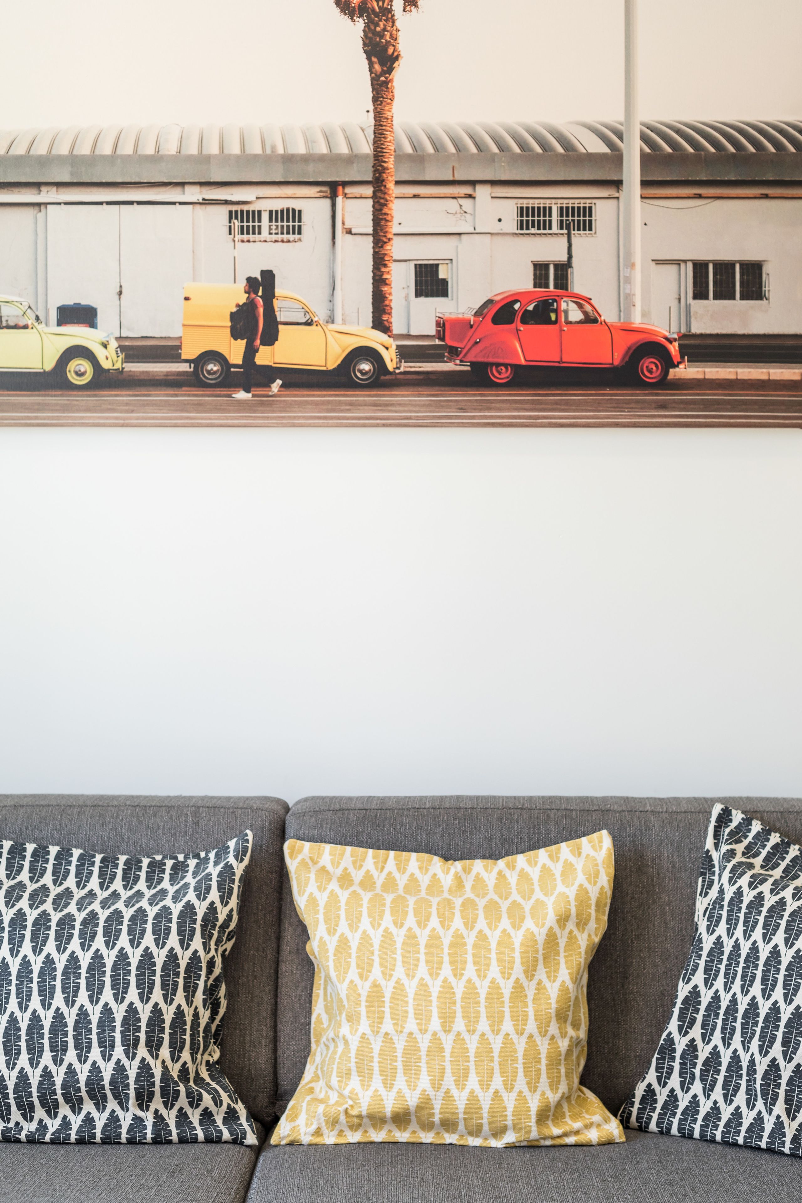 A gray sofa with patterned pillows and a view of a street with vintage cars and a palm tree.