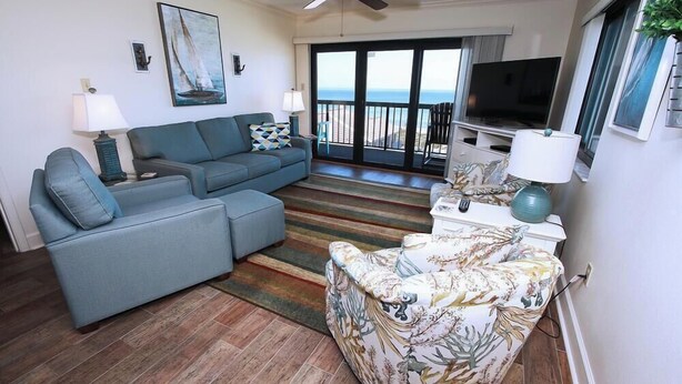 A living room with a sofa, ottoman, and patterned armchair, a television, and a view of the ocean through large windows.