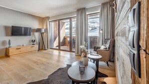 A modern living room with a flat-screen TV, wooden flooring, a sofa, a coffee table, and a view of snowy mountains through large windows. - AlpenParks Hotel & Apartment Arlberg Warth (Warth)