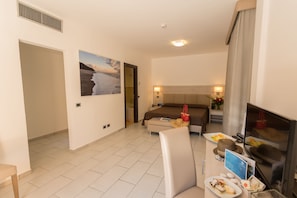 A hotel room with a bed, a TV, a dining table with food, a painting, and a bathroom visible through an open door.