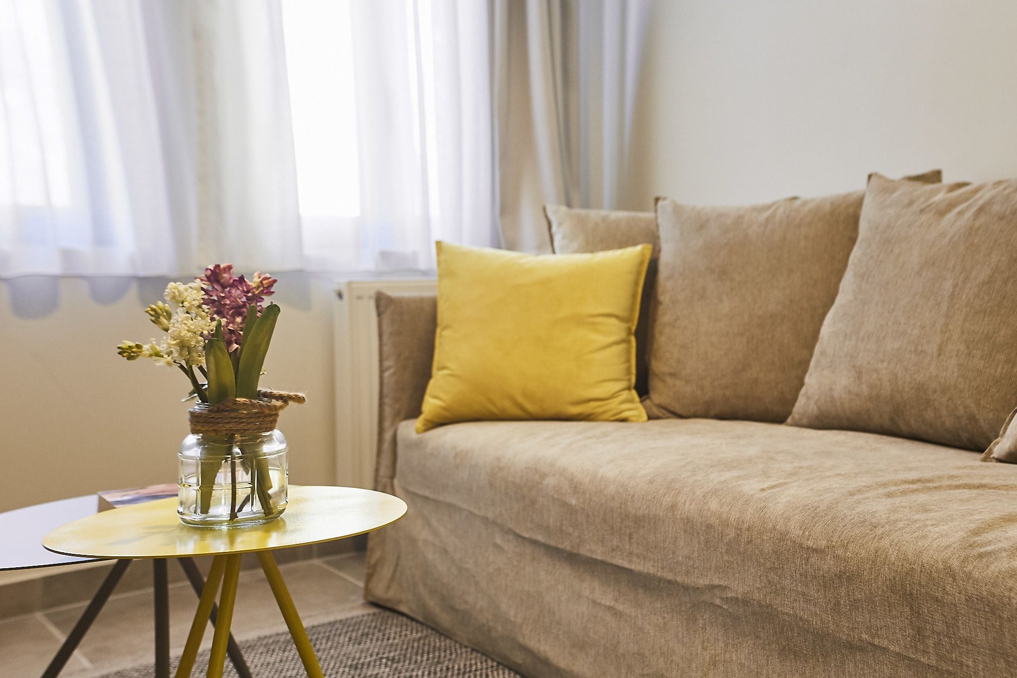 A living room with a beige sofa, a yellow cushion, and a small table with a vase of flowers.