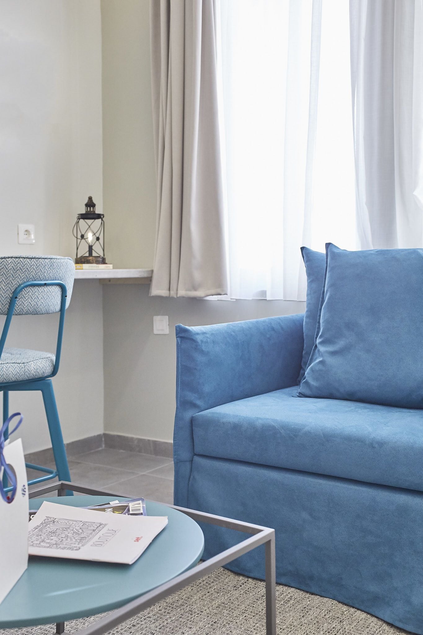 A room with a blue sofa, a blue chair, a small table with a lamp, and a window with curtains.