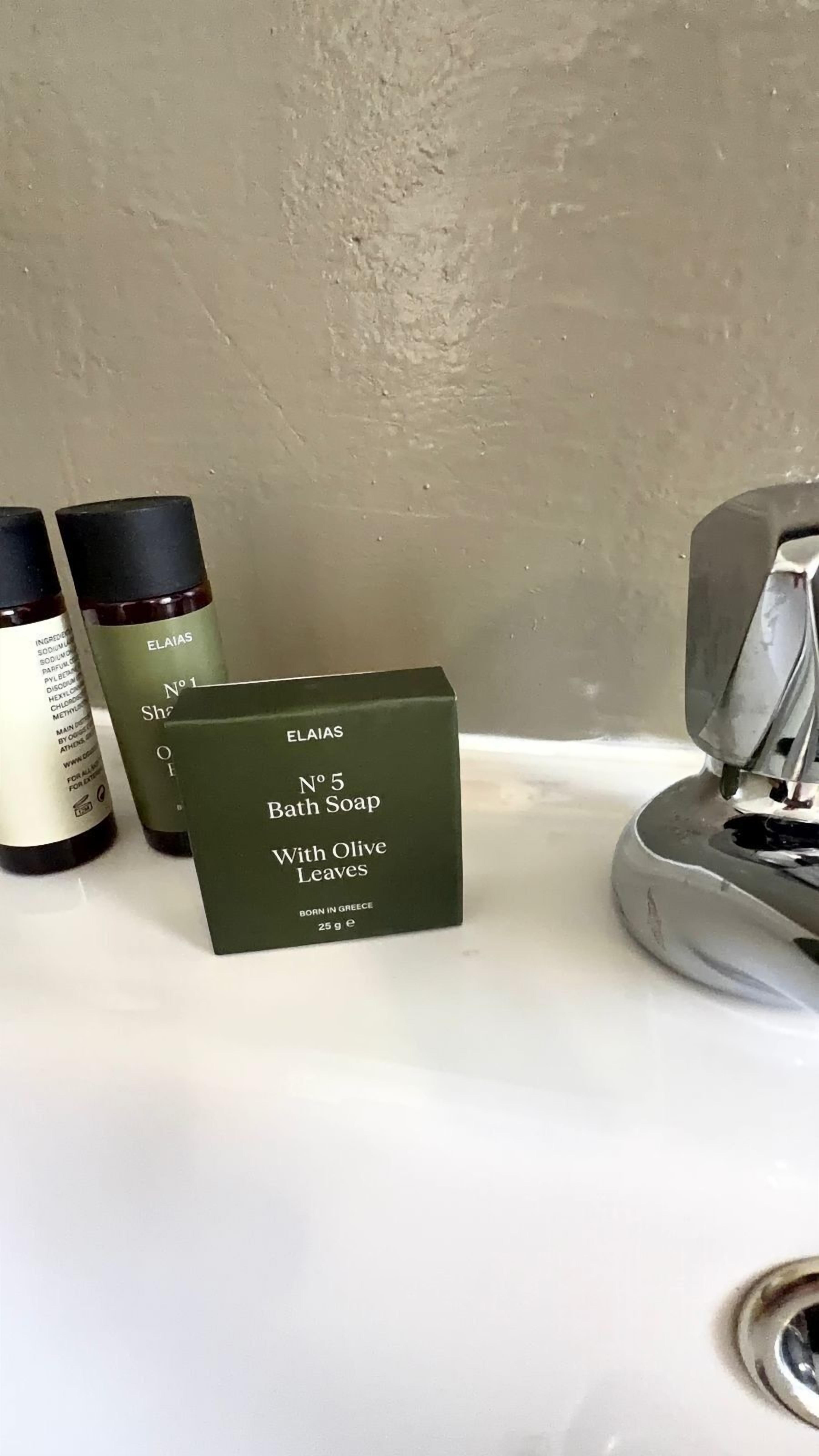Un lavamanos con una caja verde etiquetada 'Bath Soap With Olive Leaves' y dos botellas de productos de baño ELAIAS.