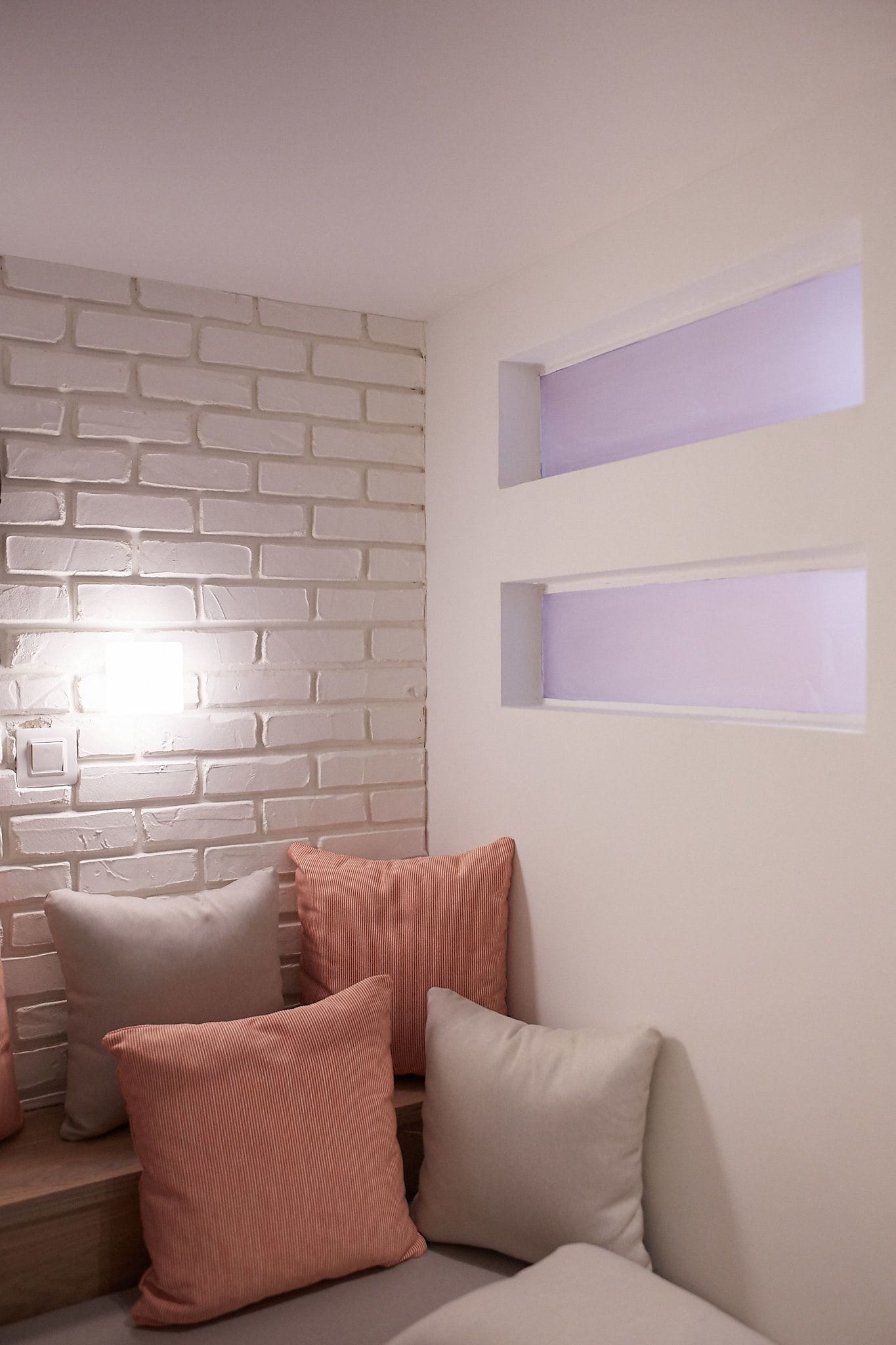 A room with a white brick wall, a window with a purple tint, and a sofa with pink and beige cushions.