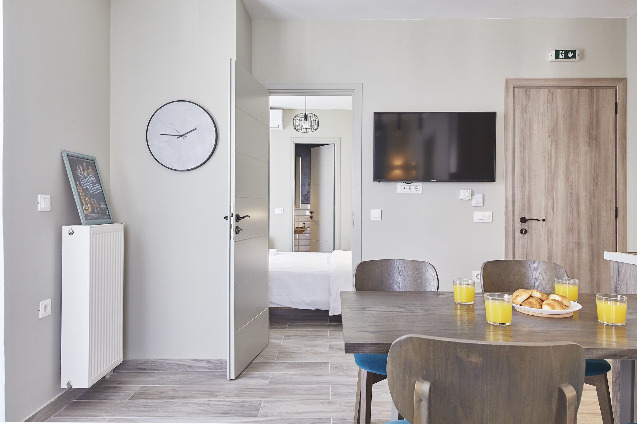 A modern hotel room with a dining area featuring a wooden table, chairs, and a plate of croissants with orange juice. A TV is mounted on the wall, and a clock is visible.