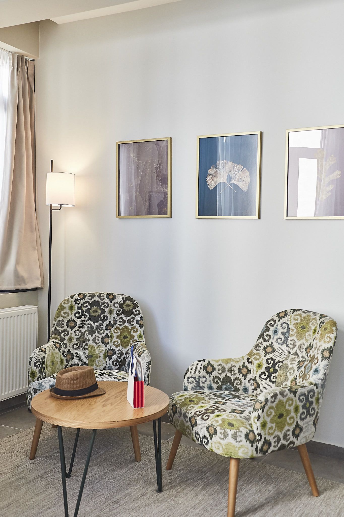 A room with two patterned armchairs, a round wooden table with a hat and a red object, and three framed artworks on the wall.