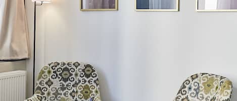 A room with two patterned armchairs, a round wooden table with a hat and a red object, and three framed artworks on the wall.