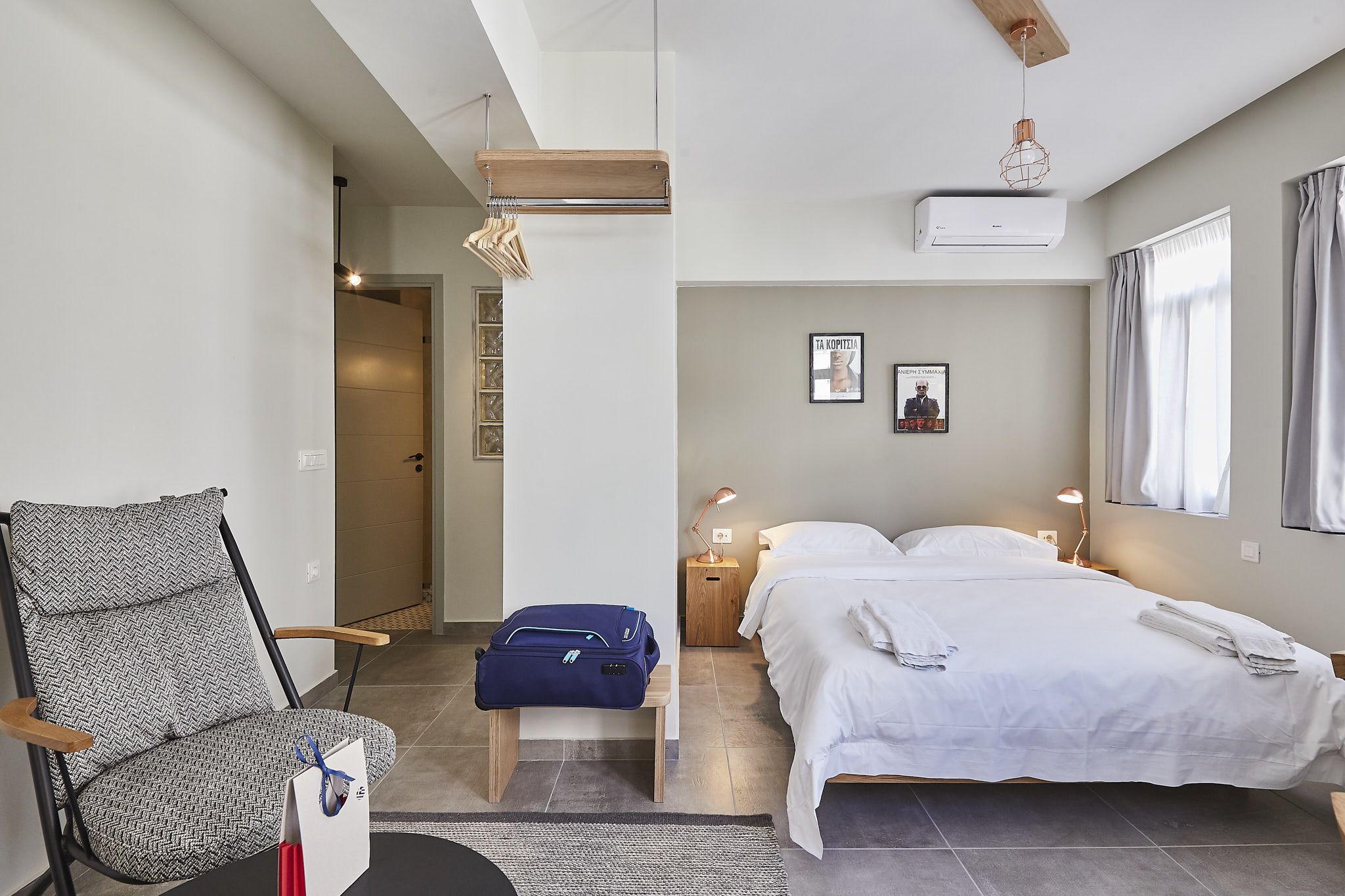 A hotel room with a bed, a chair, a suitcase, a bedside table with a lamp, and a wall-mounted air conditioner.