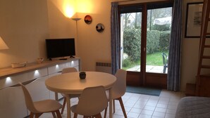 A room with a round table, four chairs, a television on a cabinet, and a view of greenery through a sliding door. - little house by the sea (Piriac-sur-Mer)