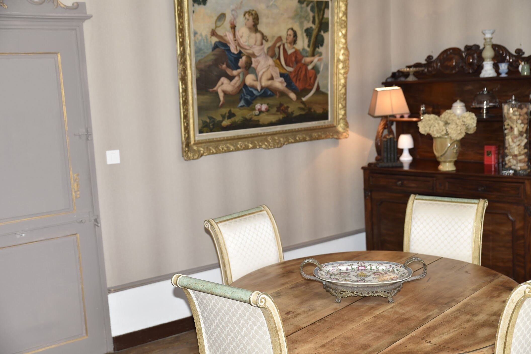 A dining table with chairs, a decorative tray, a painting on the wall, and a wooden cabinet with a lamp and flowers.