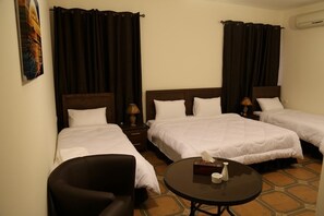 A hotel room with three single beds, a small round table, a chair, and a painting on the wall.