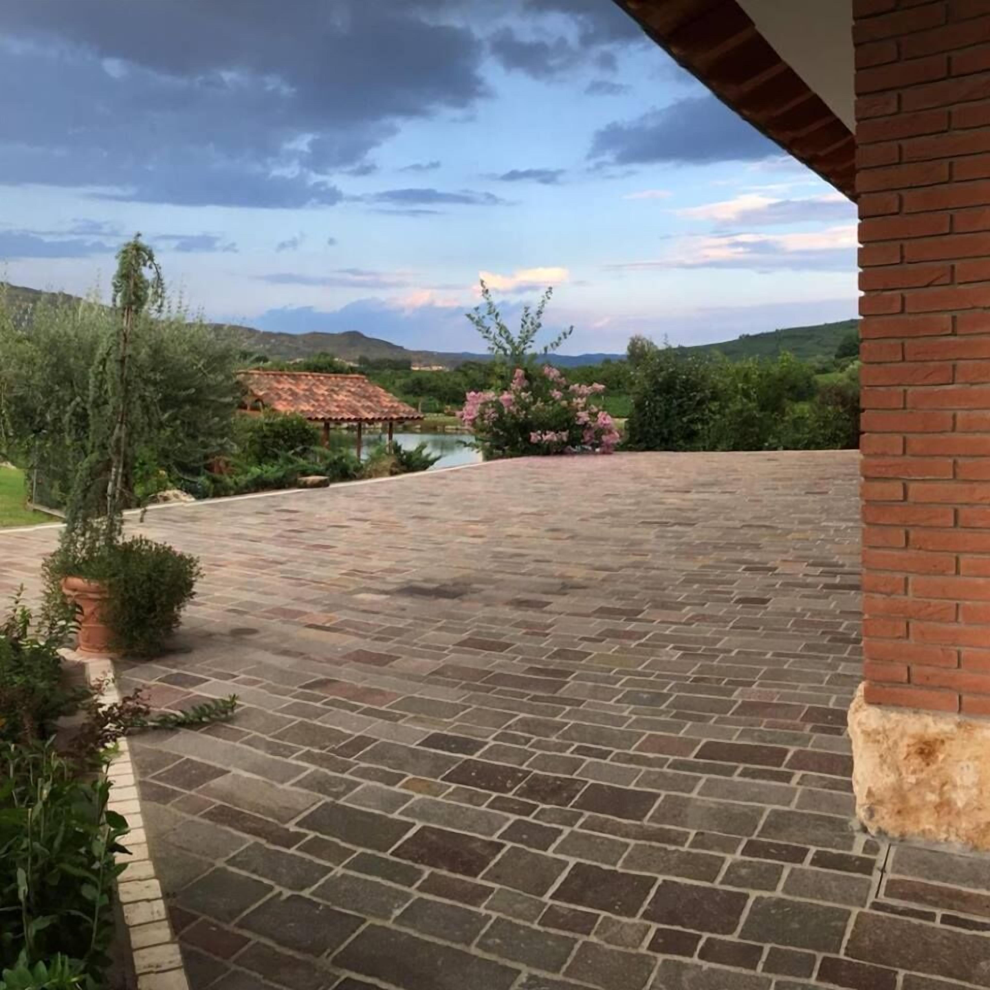 A patio area with a brick floor, surrounded by greenery and a view of hills in the distance.