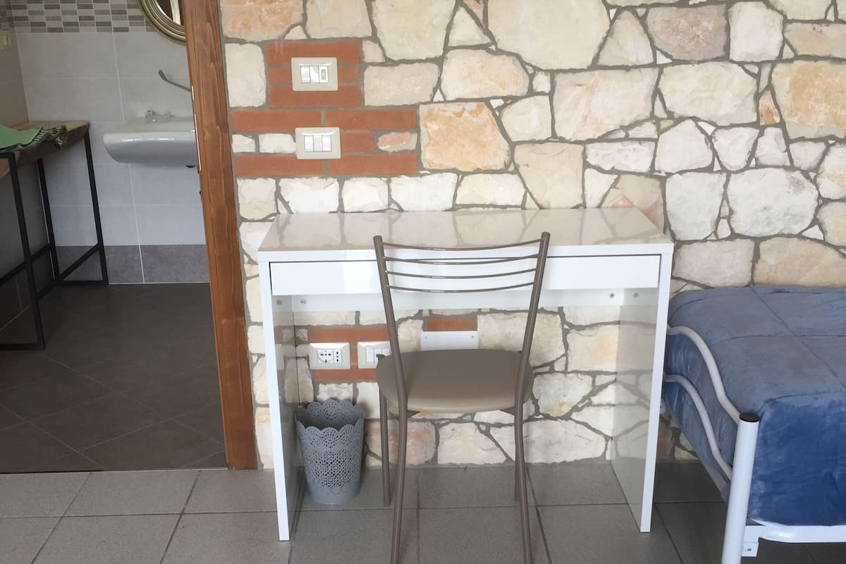A room with a stone wall, a white desk with a chair, a blue bed, and a tiled floor.