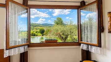 Quarto com grande janela que oferece vista para um lago, árvores e colinas.