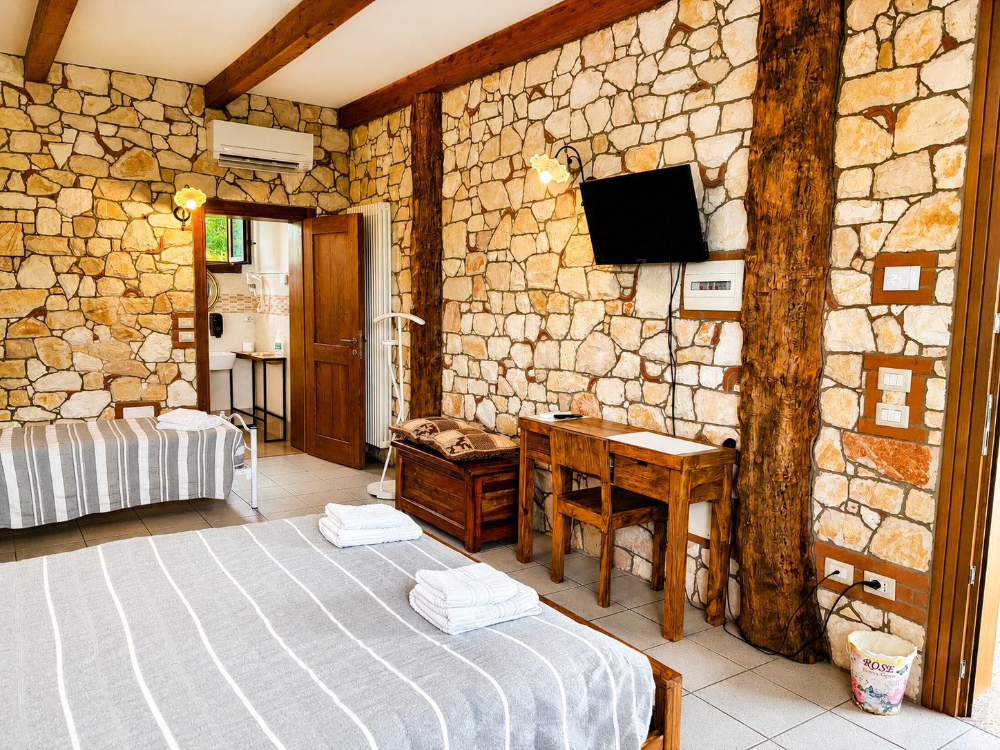 Quarto com parede de pedra, cama com roupa de cama listrada, escrivaninha de madeira, televisão fixada na parede e um balde com a etiqueta 'ROSE'.