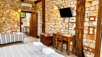 Quarto com parede de pedra, cama com roupa de cama listrada, escrivaninha de madeira, televisão fixada na parede e um balde com a etiqueta 'ROSE'.