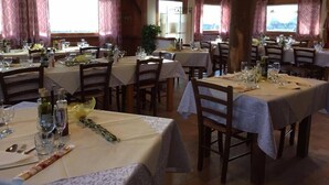 Un restaurant avec de nombreuses tables dressées pour le service, des poutres en bois au plafond et de grandes fenêtres avec des rideaux.