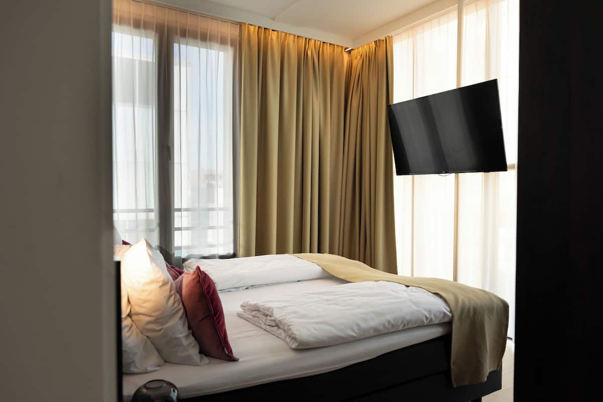 A hotel room with a bed, a flat-screen TV, and a window with curtains.