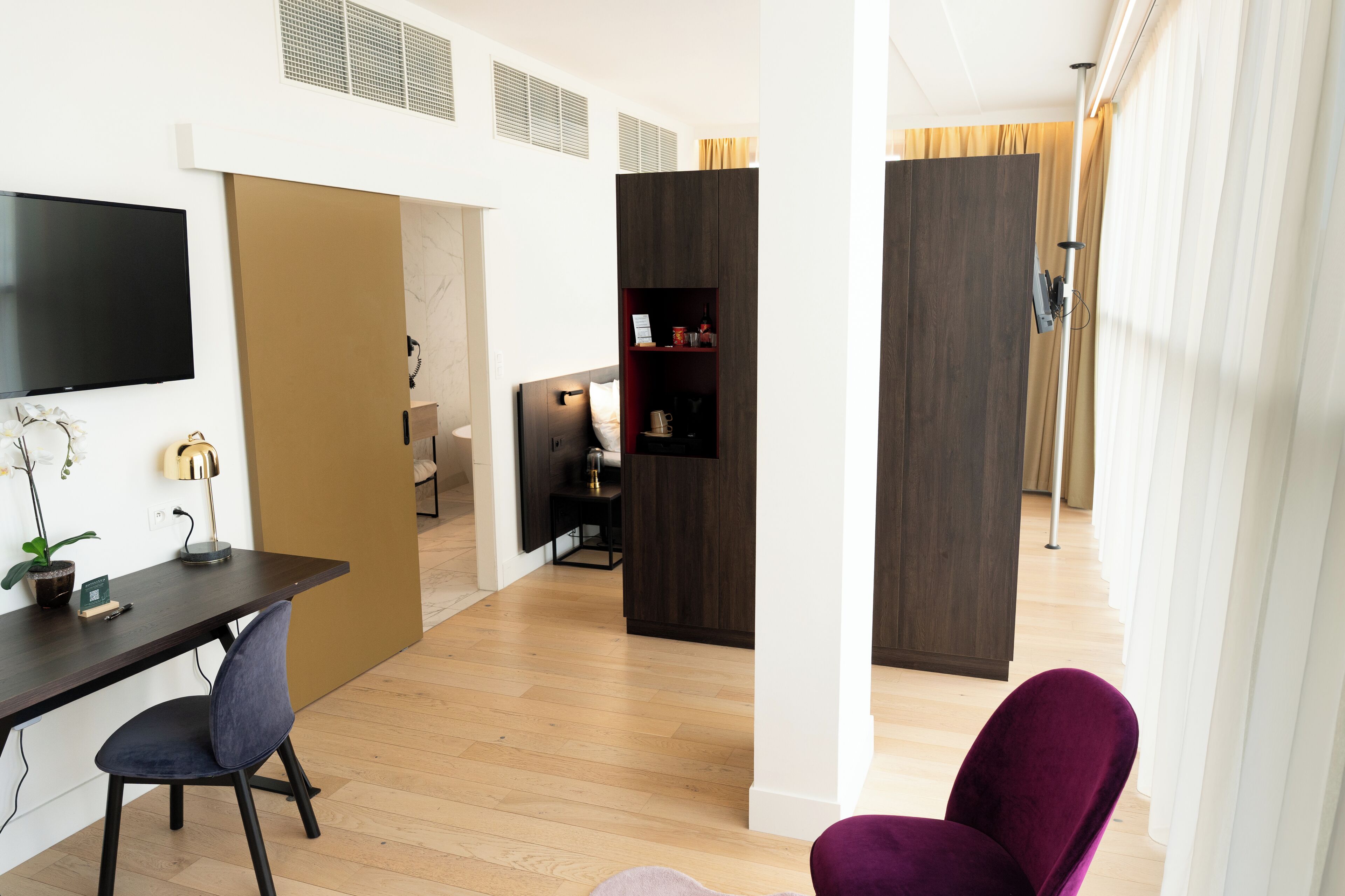 A modern hotel room with a wooden floor, a desk with a chair, a TV mounted on the wall, and a wardrobe with a mirror.