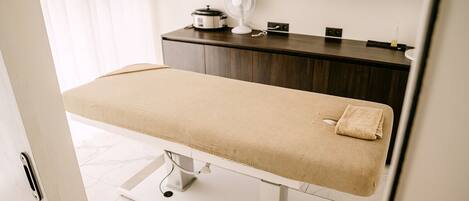 A clean, modern examination room with a white examination table, a small fan, and neatly folded towels on a shelf.